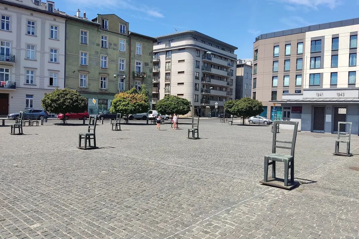 Auf einem gepflasterten Platz, gesäumt von Wohnhäusern unter blauem Himmel, stehen große leere Stühle.