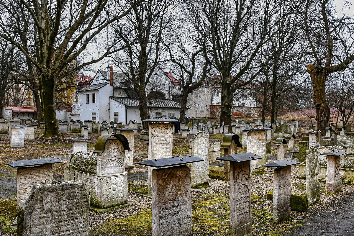 Zwietrzałe kamienne macewy z wyrytymi hebrajskimi inskrypcjami stoją wśród bezlistnych drzew na starym cmentarzu Remuh, a w tle widać białe budynki.